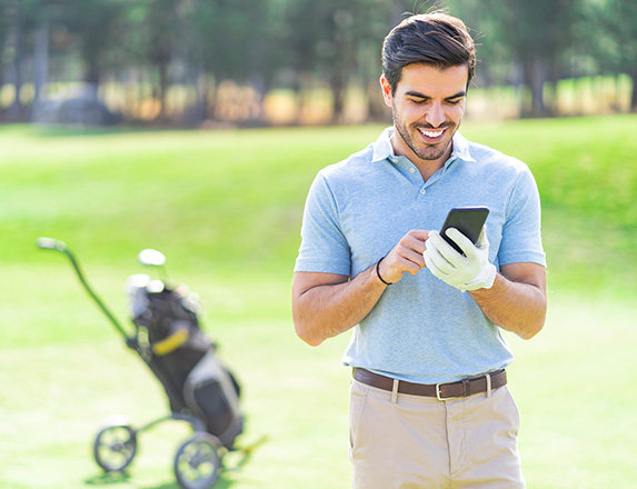 a smiling man looking down at his cell phone while on the golf course