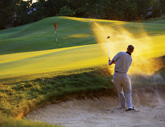 a man hitting a golf ball in the sand with the sun setting to the right