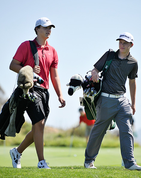two young golfers walking on the course with their bags over their right shoulders