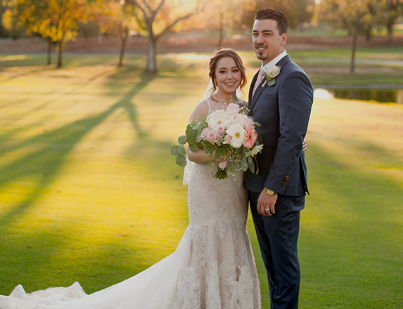 a smiling bride and groom posing for a photo on the golf course with the sun setting just behind them