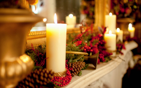 Lit up candles with holly berries & pine cones on top of fireplace