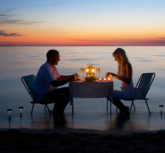 man and woman having a romantic candlelit dinner on the water with the sunset in the background