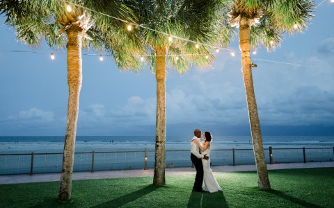 married couple kissing underneath palm trees with ocean in background