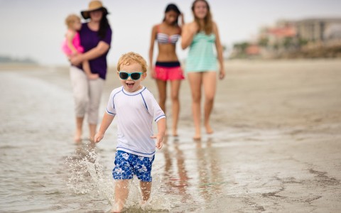 young boy in swimsuit splashing at waters edge while family walks a little behind him in sand