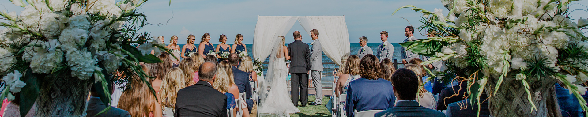 Wedding ceremony with guests watching bride and groom exchange vows under fabric draped arch with an ocean backdrop
