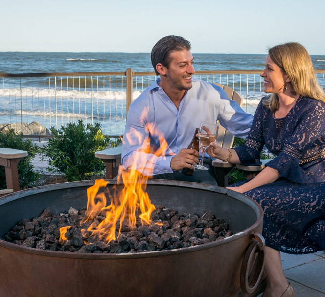 well dressed man and woman sitting drinking wine in front of an outdoor fire pit on the patio along the ocean shore