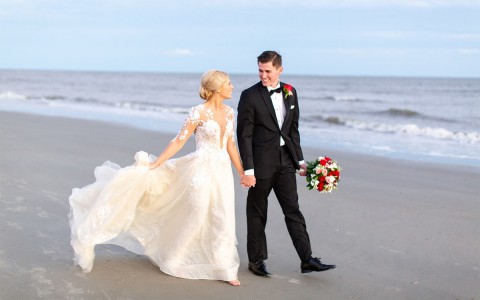 bride and groom holding hands and looking at each other while walking on the beach