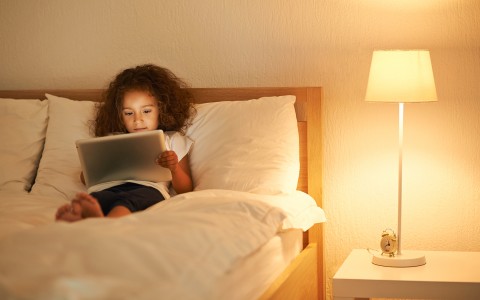 Kid lying down in the bed while she is looking at the Ipad