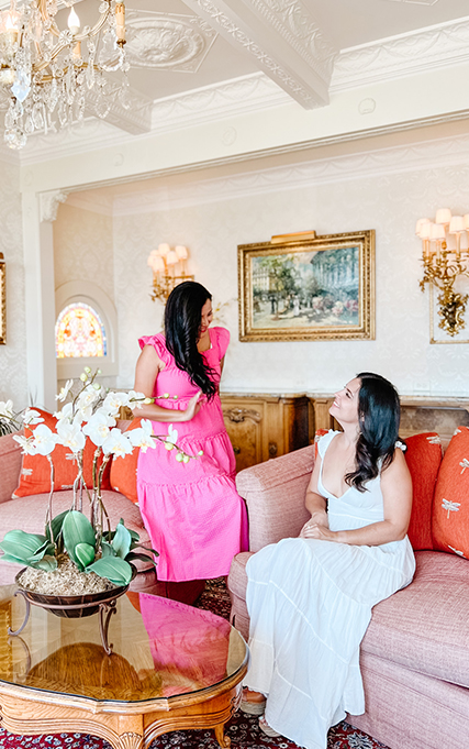 Two girls with long black hair wearing dresses and smiling at each other in a white room.