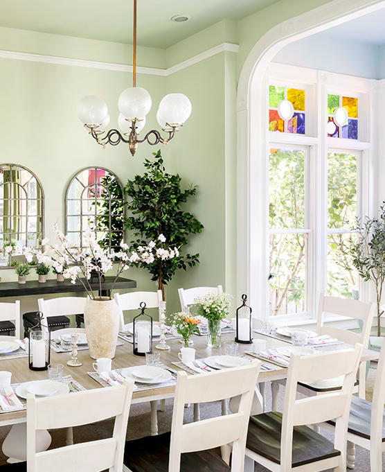 A long dining room table with white chairs and a chandelier hanging above with plants and flowers during the day.