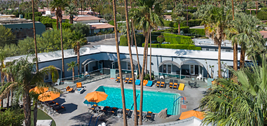 A drone shot of a pool surrounded by tall palm trees and an l-shaped building behind it.