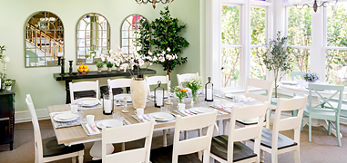A light green dining room with a large white rectangle table and the sun shining in from the windows.