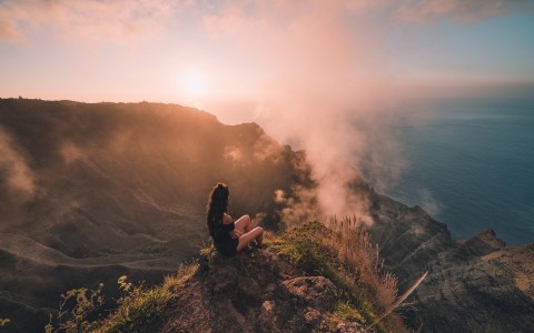 woman sitting at the top of a mountain looking at the ocean during sunset