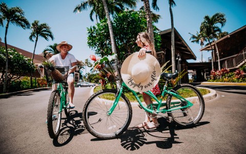 woman holding a sun hat while standing next to a bike and a man riding a bike next to her