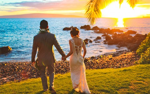 a bride and groom holding hands and walking towards the ocean while the sun is setting