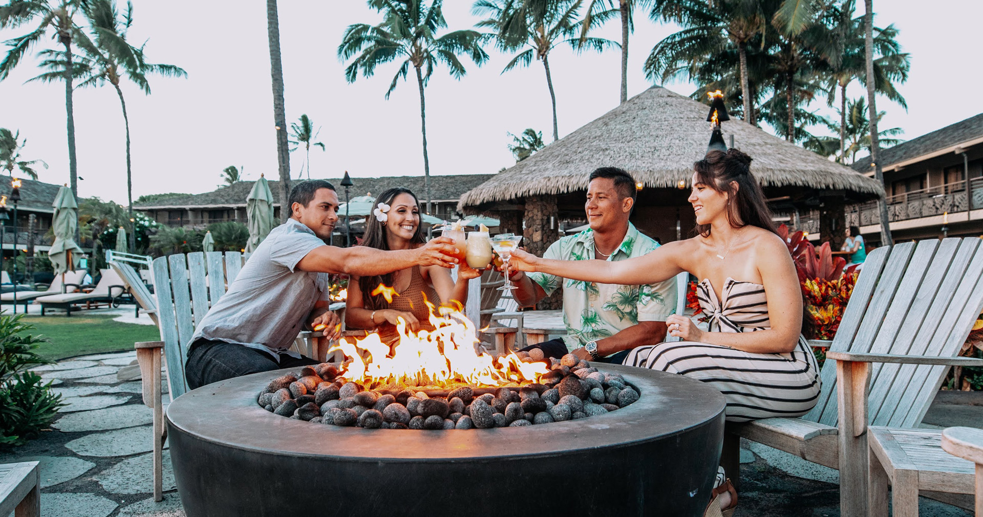group of people sitting around a firepit clinking their drinks
