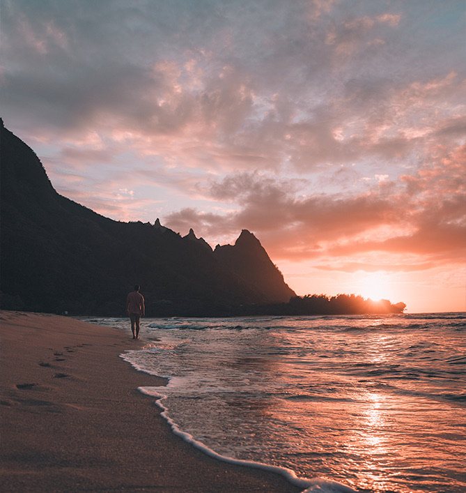 view of a man walking on the shore with the mountains in the background while the sun is setting