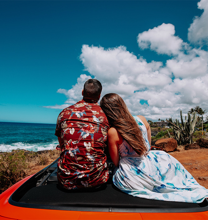 a woman resting her head on a mans shoulder while sitting in a red convertible looking at the ocean