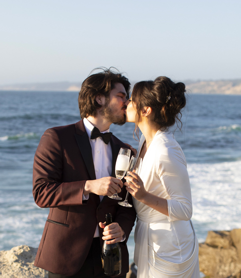 couple in bridal wear kissing with champagne glasses by ocean