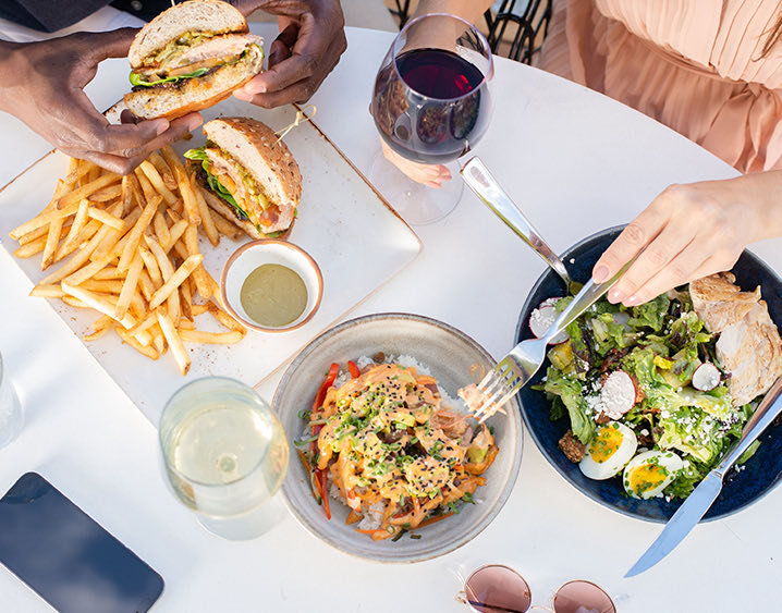 people eating a salad, chicken sandwich with french fries, and another dish with wine