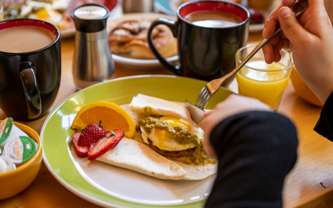 a person cutting a tortilla on a plate with fruit by two cups of coffee and a glass of orange juice
