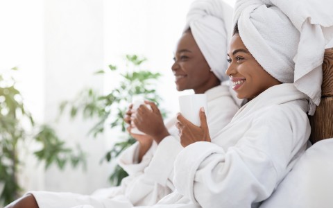two women in robes and towels wrapped around their heads holding cups of coffee and smiling