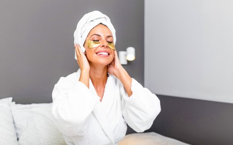 woman sitting with under eye patches in a robe and a towel wrapped around her head smiling