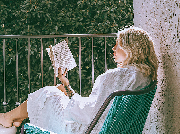 woman sitting on blue patio chair wearing white robe and reading a book