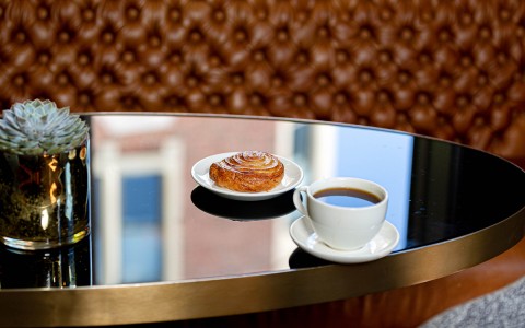 cup of coffe placed on table with a pastry on a plate behind it