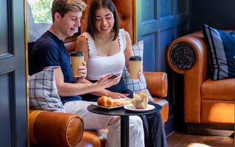two people sitting and holding coffee to go cups looking at a phone beside a small table with a muffin and pastry on it