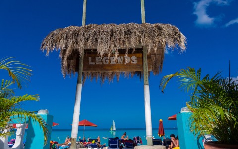 Legends tiki bar sign entrance with people relaxing on the sand with clear blue water in background