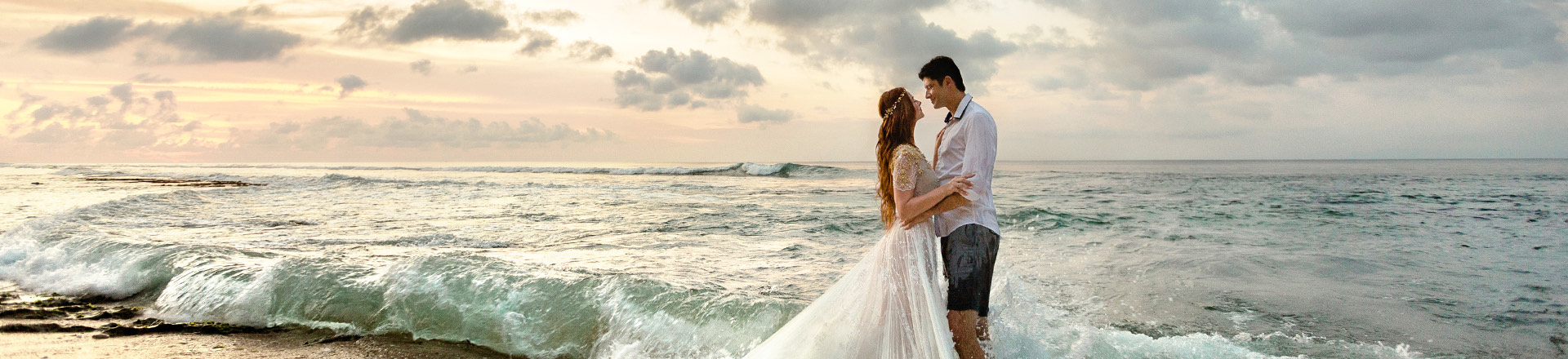bride and groom embraced in each other's arms standing on edge of beach with waves crashing at shore, romantic cloud sunsetting in distance