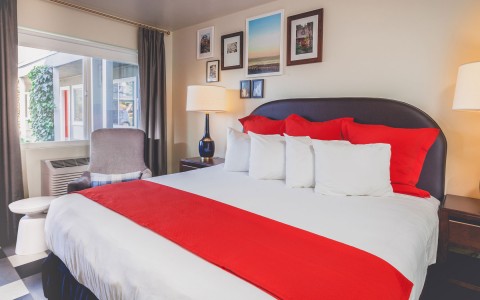 kind bed with white sheets and bright red pops of color from pillows and through blanket. Next to large window with ac unit. the wall decorated with collage of images