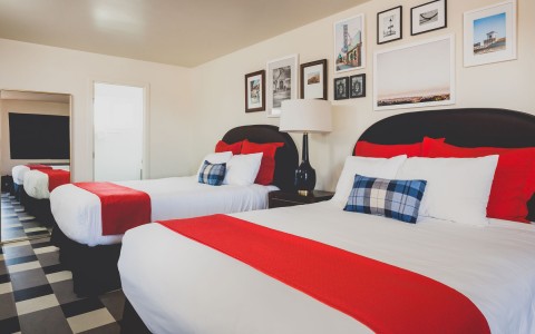 2 queens beds with white sheets and bright red pops of color from pillows and through blanket. leading to bathroom door. the wall decorated with collage of images