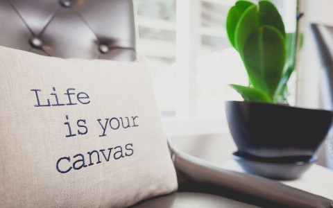 pillow with life is your canvas written on it. sitting on leather tufted chair next to table with plant