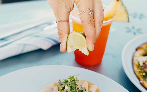 plate with 3 tacos on it, a hand is squeezing a lime ontop of the tacos. Cocktail in the background on the table