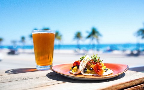 outside table with 2 tacos and a glass of beer with the beach behind it