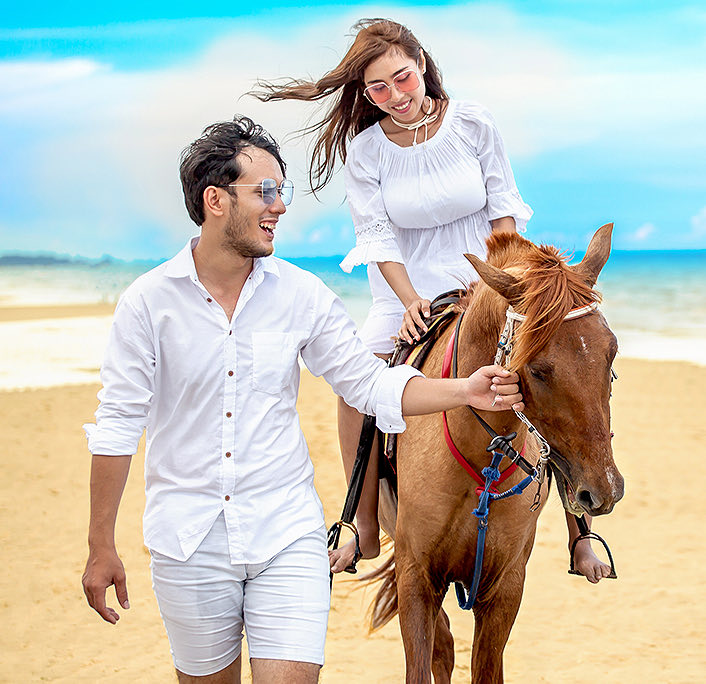 A woman horseback riding at the beach with his boyfriend next to her