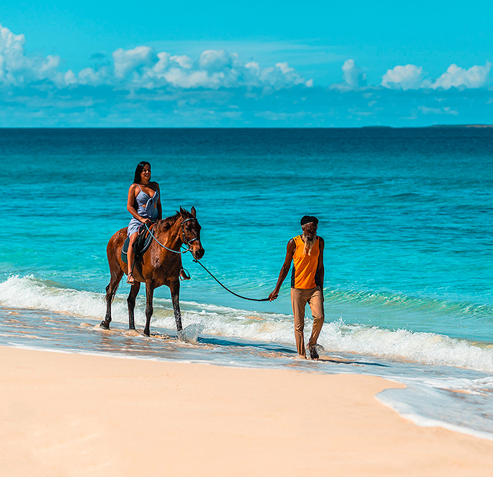 A man carrying a horse with a woman on top of it next to the sea