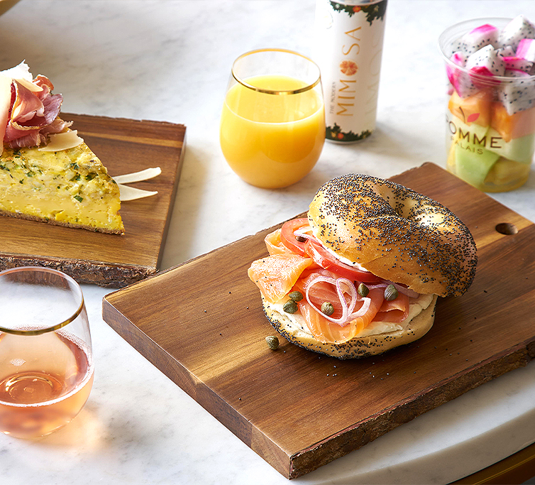 poppy seed bagel sandwich sitting on a wooden plate with a frittata orange juice and a fruit cup next to it  