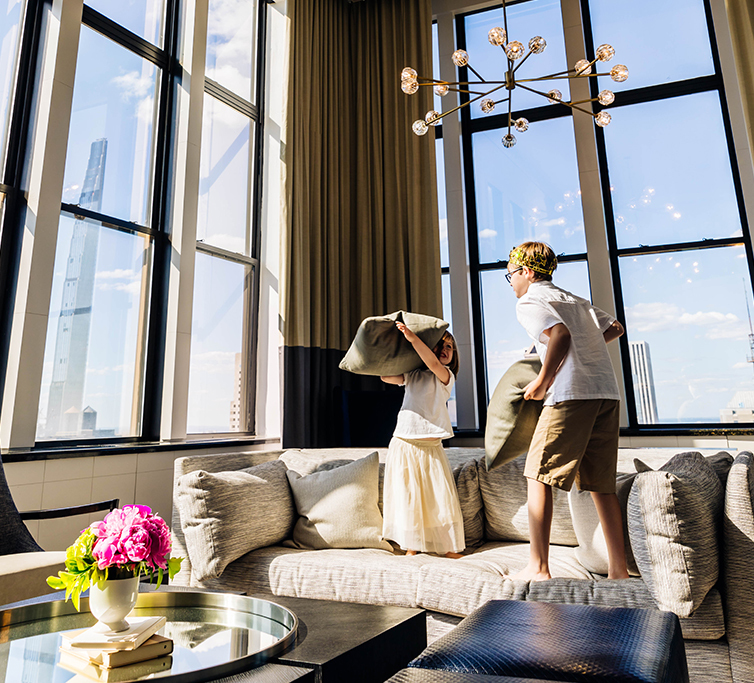 family values package image of two siblings, one boy and one girl, on the couch of the royal suite having a pillow fight. there are tall ceiling to floor windows and curtains in the background letting in warm lighting that make the flowers on the living r