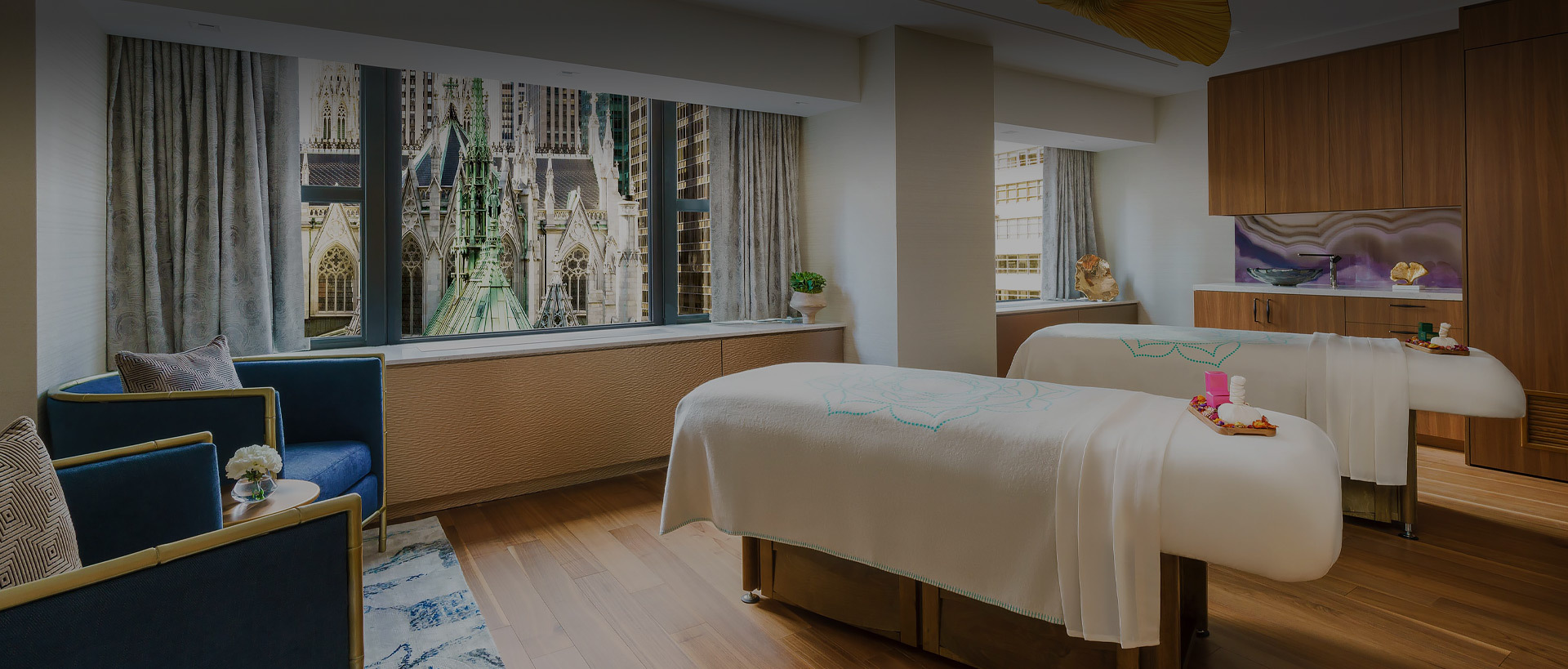 two massage tables with white sheets and flowers and candles. there are two blue chairs in the massage room with a wall to wall window to views of new york city