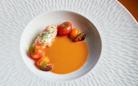 A small bowl of tomato soup on a white plate with small accents of tomates and lobster
