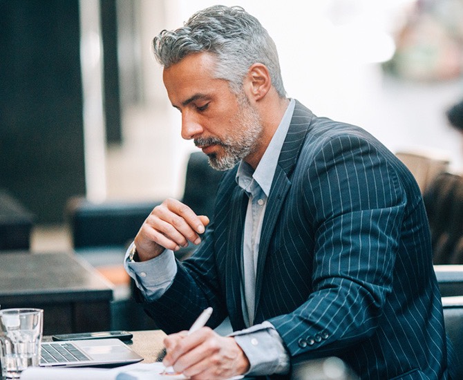 Man in suit writing notes and sitting at a table with his laptop