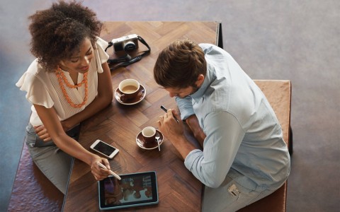 two people at a cafe table drinking a coffee cup and looking at a tablet 