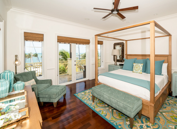 Room with King bed & large wooden frame, mint nightstands & lounging chair next to balcony