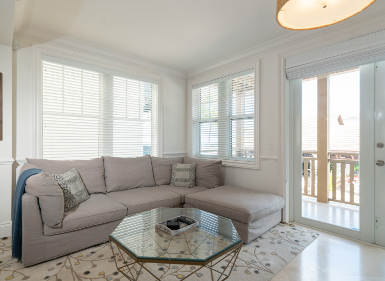 In room living space with taupe sectional, beige rug & glass table next to door leading to balcony