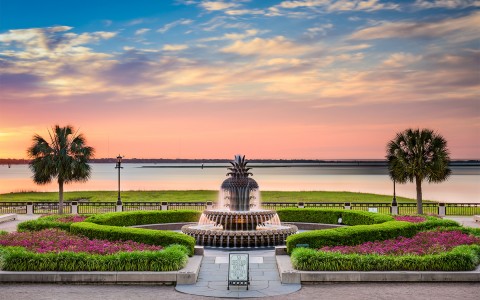 The sun setting over the water with a fountain and colorful plants.