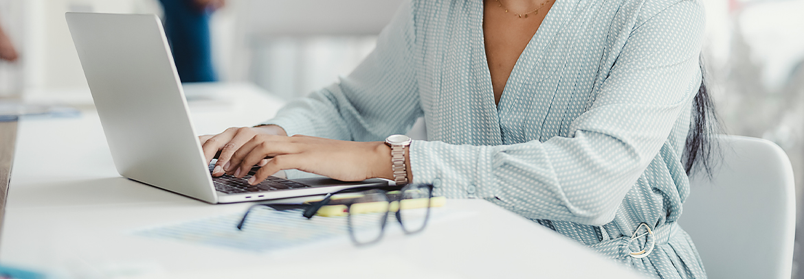 A woman typing on a laptop with her glasses next to it.