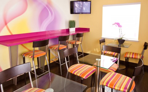 dining area contains small glass tables with 2 chairs and a wall of cafe bar seating. The room is decorated with bright wall paper, chairs and accents.